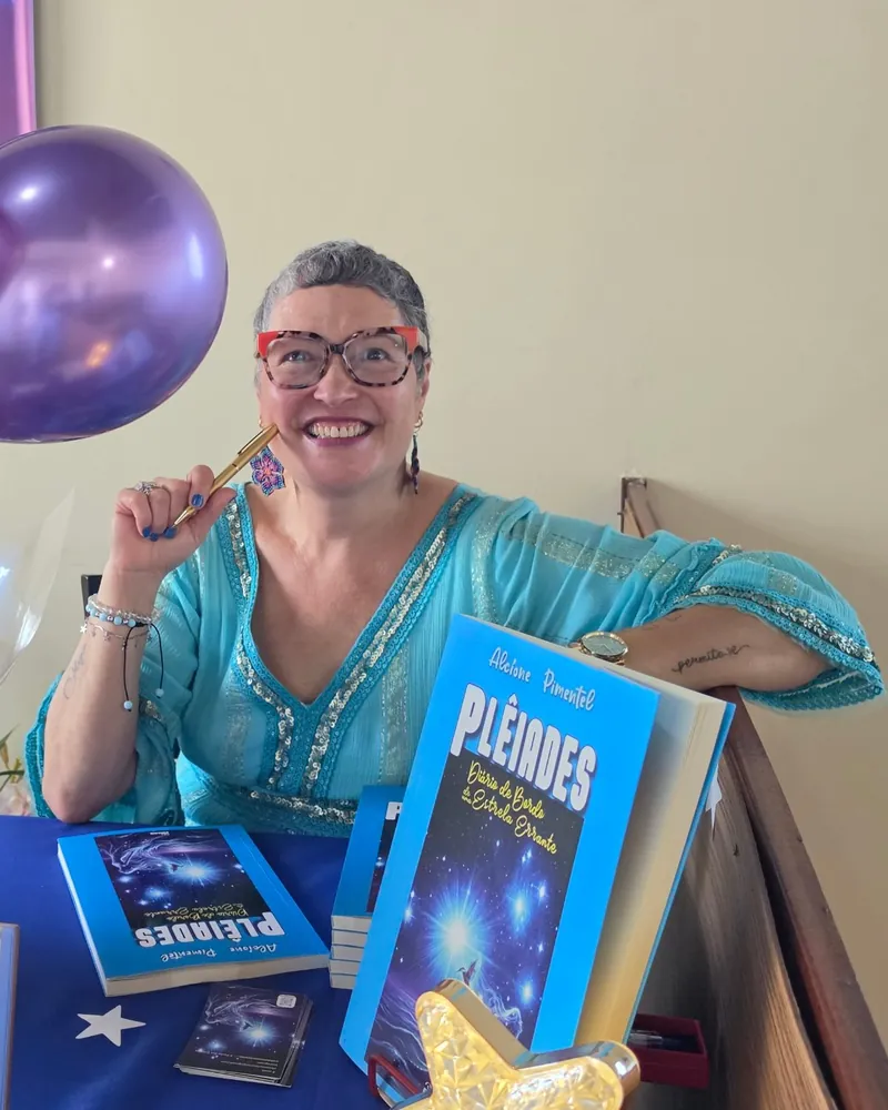 Alcíone Pimentel sorrindo, de caneta na mão, na mesa de autógrafos do lançamento de Plêiades